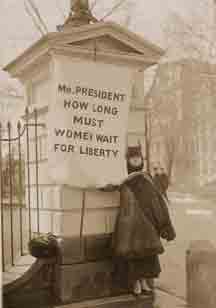 Femal protestor stands before gate of the White House with banner that says, "Mr. President, How long must women wait for liberty"
